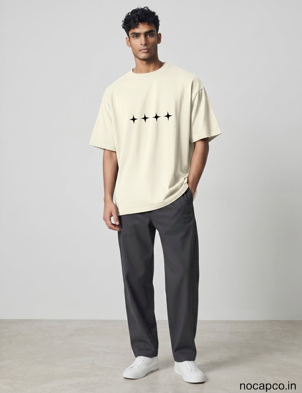 No Cap Co cream oversized cotton t-shirt with four black star graphic print on chest, worn by a man with dark hair and grey trousers.
